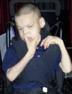 Photo of Jesus Hibbard, a boy in a wheelchair. He has light-tan skin and black hair cut short in a buzz cut. He looks somewhat confused. One of his hands is up in front of his face. He is wearing a safety belt over his blue collared shirt.
