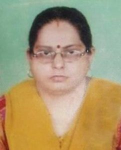 Photo of Swarna Kapoor. She has fair skin, a round face, and dark brown hair neatly pulled back. She is wearing glasses and has a dark-red bindi on her forehead. Her dress is red with a wide yellow collar.