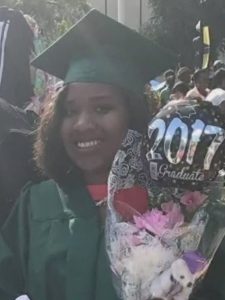 Photo of Kiara LaSalle, a teenage girl in a green graduation cap and gown, smiling broadly. She has curly brown hair and brown skin and is carrying a bunch of flowers and a balloon that says "2017 graduate".