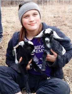 Photo of Savannah Leckie, a teenage girl with fair skin and light-brown hair, wearing a knit cap and sweatshirt. She is holding two black-and-white goat kids.