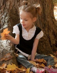 Photo: Small fair-skinned girl seated at the base of a tree, her blond hair in ponytails, holding and looking at a leaf. She is wearing a frilly white blouse, black vest, jeans, and pink sneakers.