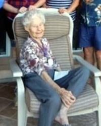 Photo of Vivian Nelson, an elderly woman with pale skin and short curly white hair. She is thin, wearing a floral blouse and blue slacks, and sitting in a big beige chair with one leg propped up on the chair.