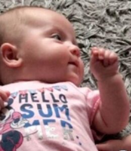 Photo: A baby lying on her back; she has light skin and is bald, and is wearing a pink shirt that reads Hello Summer.