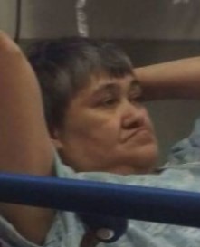Photo of an older woman with fair skin and gray hair cut in a businesslike style, lying back in a hospital bed with her head propped on her hands.
