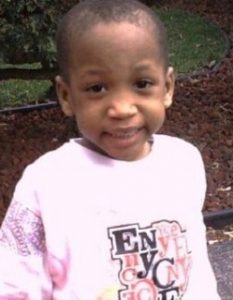 Photo of Davarion Davis, a boy with brown skin and black hair in a buzz cut, photographed outdoors, smiling at the camera. He is wearing a pink sweatshirt with a logo printed on the front.