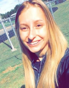 Photo of Kayla Hensley, a teen girl with long, straight blond hair, fair skin, and braces on her teeth. Behind her is a green lawn and a fence. She is wearing a dark blue top and smiling.