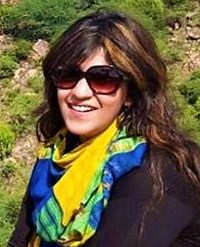 Photo of Sana Iqbal, a young woman with light skin and wavy dark-brown hair. She is wearing a big pair of sunglasses, a colorful blue and yellow scarf, and a jacket. The photo is taken outdoors.
