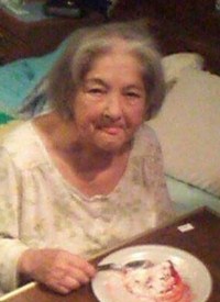 Photo of Beverly Nickerson, an elderly woman with shaggy gray hair and pale skin, wearing a nightshirt and sitting on the edge of a bed, eating from a plate on a bedside table. The photo has caught her in the middle of licking her lips.