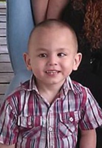 Photo of Jordan Rodriguez, a small boy with light skin and brown eyes. He is smiling. He has a lazy eye. His brown hair is cut very short, and he is wearing a red and white checked shirt.