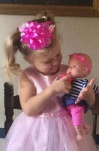 Photo of Mariah Woods, a toddler girl with fair skin and blonde hair in two ponytails. She is wearing a pink formal dress and has a big pink gift ribbon glued to her hair; she is pretending to feed a baby doll.