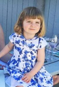 Photo: A toddler girl, sitting, in a white dress with blue flowers. She has shoulder-length blond hair and fair skin.