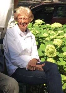 Photo of Barbara Wilmes, a light-skinned, white-haired elderly woman wearing a white shirt and dark pants, smiling as she sits on a bench in front of a flower bush.