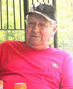 Photo of Douglas Fischer, an elderly man, somewhat chubby, wearing a baseball cap and T-shirt and leaning back against a metal fence, looking contemplatively into the middle distance through wire-rimmed glasses.