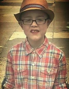 Photo of Ethan Okula, a small boy with fair skin. He is wearing a straw trilby, dark-rimmed glasses, and a flannel shirt.