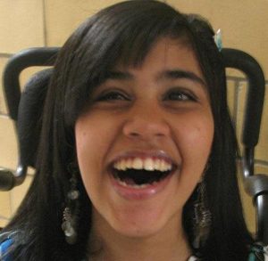 Photo of Qyzra Walji, a young woman with straight brown hair and tan skin, smiling broadly. A wheelchair head rest is visible behind her head.