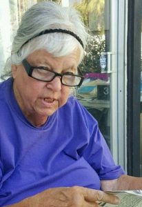 Photo of Lerae Bush, an elderly woman with fair skin and white hair held back in a headband. She is wearing reading glasses and holding a newspaper.