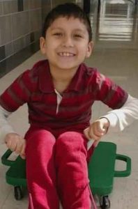 Photo of Abraham Cardenas, a little boy with olive skin and dark hair, smiling for the camera. He is wearing a red striped shirt and red pants, and is sitting on a scooter board.