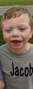 Photo of Jacob Edwards, a small boy with fair skin and blond hair, smiling for the camera. He is wearing a gray T-shirt that says "Jacob".