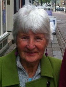 Photo of Betty Lyons, an elderly woman with short white hair and freckled skin, wearing a green coat and purple drop earrings.