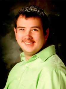 Portrait photo of Cody Nachtrab, a teenager with dark hair and light skin and a wispy mustache. He is smiling for the camera. He is wearing a pale-green dress shirt.