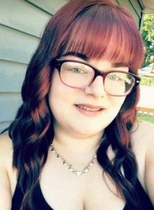 Photo of Destiny Rollar. She has pale skin and hair dyed auburn. She is wearing big glasses, a black strappy dress, and a gold heart necklace.