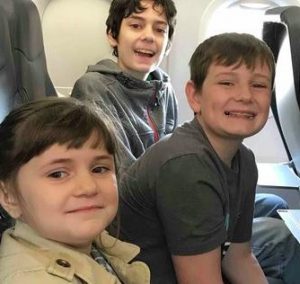 Photo of Tyler, Aidan, and Arianna Talmage. They are children with light-olive skin and brown hair, photographed sitting in a row of airplane seats and smiling for the camera.