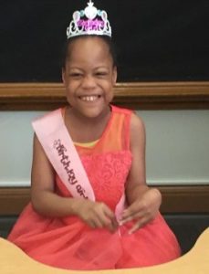 Photo of Heaven Watkins, a small girl with brown skin and black hair pulled back under a silver crown. She is wearing a coral-colored dress and a sash that says "Birthday Girl". Her face is scrunched up in a wide smile.