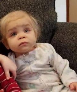 Photo of a baby with pale skin and wispy, light-blonde hair, wearing pajamas with a zipper up the front. She has a puzzled, slightly worried expression on her face.