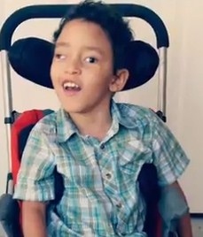 Photo of a young boy with light-brown skin and curly black hair sitting in a wheelchair with a headrest. He is smiling for the camera.