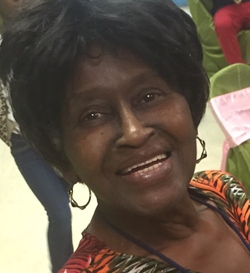 Photo of a middle-aged woman with dark brown skin and short, straight black hair. She is smiling at the camera; her eyes are twinkling. She is wearing big hoop earrings and an orange-and-green print top.