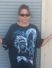 Photo of Dawn Liebig, a middle-aged woman with tanned skin and brown hair in a messy ponytail. She is wearing sunglasses and a T-shirt printed with a skeleton in a feathered war bonnet and a wolf howling at the moon. She has her hands upturned in an "I don't know" gesture.