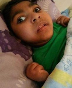 Photo of a boy lying in bed, covered with a blanket. He has dark-brown eyes, brown skin, and dark hair. His hands are balled into fists.