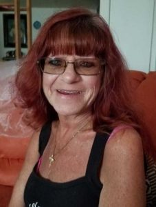 Photo of a middle-aged woman with dyed red hair, wearing a black tank top and a gold cross necklace. Her skin is freckled, and she wears sunglasses.