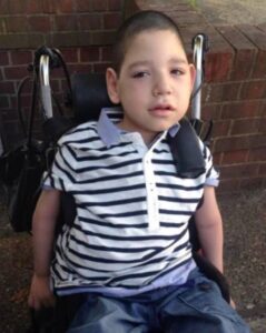 Photo of a boy sitting in a wheelchair; he has light skin, dark brown hair, and a small head; he is wearing a blue and white striped polo shirt and jeans.
