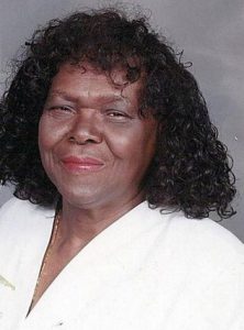 Photo of Mary Smith, a heavyset woman with dark-brown skin and shoulder-length hair in ringlets. She is wearing lipstick and a white blouse, and squinting at the camera.