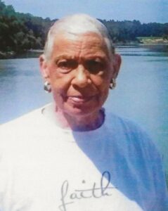 Photo of an older woman with very short gray hair, dark skin, and white shirt, photographed outdoors.