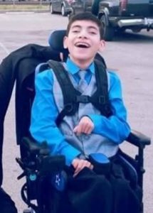 Photo of a teenage boy in a power chair, wearing a blue shirt and tie. He is smiling broadly. His dark hair is cut short.