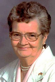 Portrait photo of Evelyn Bailey, an elderly woman wearing a white suit jacket and a string of pearls. Her salt-and-pepper hair is permed and cut short. She has pale skin and is wearing large glasses and a flag lapel pin. She is smiling for the camera.