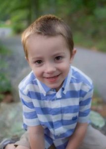 Photo of a small boy sitting on the ground, leaning toward the camera and smiling. He has fair skin and short blond hair, and is wearing a blue and white striped shirt.