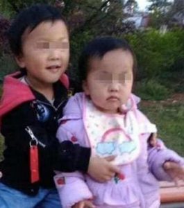 Photo of two Chinese toddlers, a boy in a black and red coat, and a girl in a purple coat. The boy has his arms around the girl. Their eyes are pixelated to keep them anonymous.