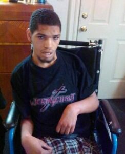 Photo of a young man sitting in a wheelchair. He has short curly black hair and beard.