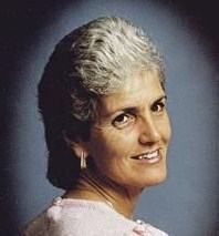 Portrait photo of an older woman wearing a pink blouse and pearl drop earrings. Her gray hair is cut short.