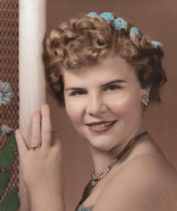 Colorized black-and-white photo of a young woman with curly hair, fair skin, and blue flowers in her hair, smiling for the camera. She has an engagement ring on her finger.