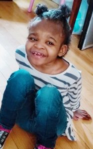 Photo of Zha-Nae Wilkerson, a small African-American girl sitting on the floor, knees pulled up to her chest, smiling for the camera.