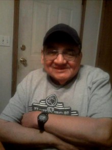 Photo of Elvis Dry, an older man with fair skin and a large nose, grinning at the camera with his arms folded. He is wearing glasses, a baseball cap, a sweater, and a wristwatch.