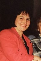 Photo: A middle-aged woman with dark hair and light skin, in a soft orange suit, smiling for the camera.