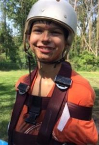 Photo of a smiling boy in a helmet. His arms are tied to his side with straps. He has fair skin and dark hair and is wearing an orange shirt.
