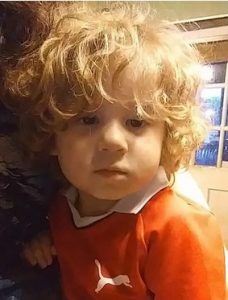 Photo of Dante Mullinix, a small boy with a mop of curly blond hair, wearing an orange polo shirt and looking at the camera.