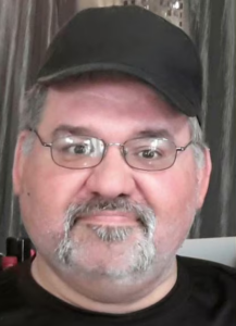 Photo of a round-faced, middle-aged man with light skin and a neat salt-and-pepper mustache and short-cropped beard. He is wearing a black baseball cap.