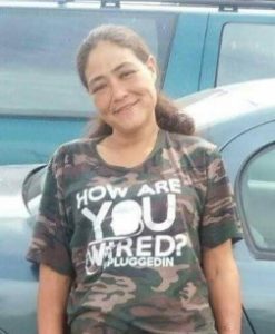 Photo of a young woman with bushy brown hair and fair skin, leaning against a car, wearing a printed camo T-shirt that says, 